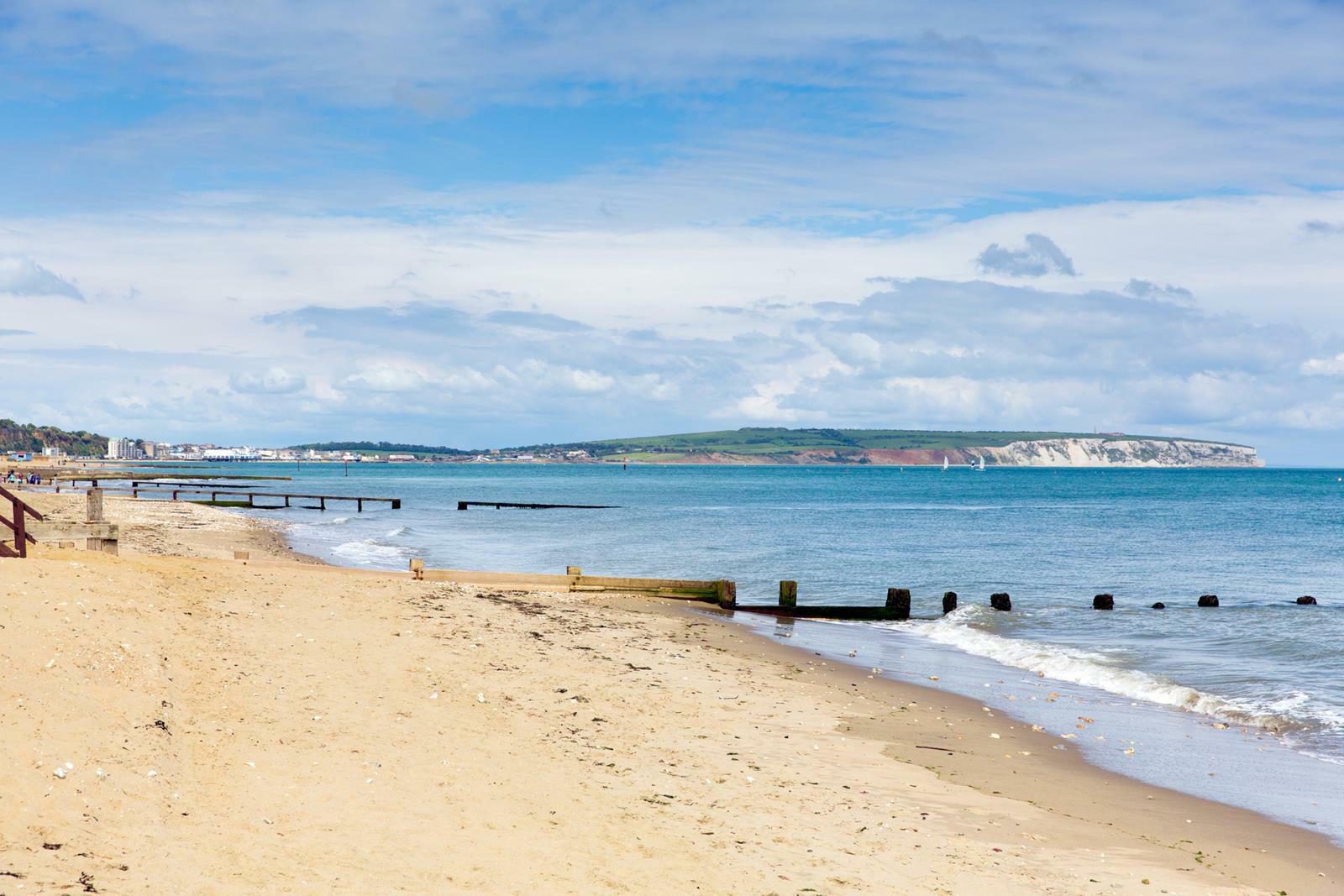 A tranquil sandy beach with gentle waves rolling onto the shore. Wooden groynes extend into the sea, and in the distance, a green, hilly coastline stretches under a partly cloudy sky. Some buildings and structures are visible far off to the left.