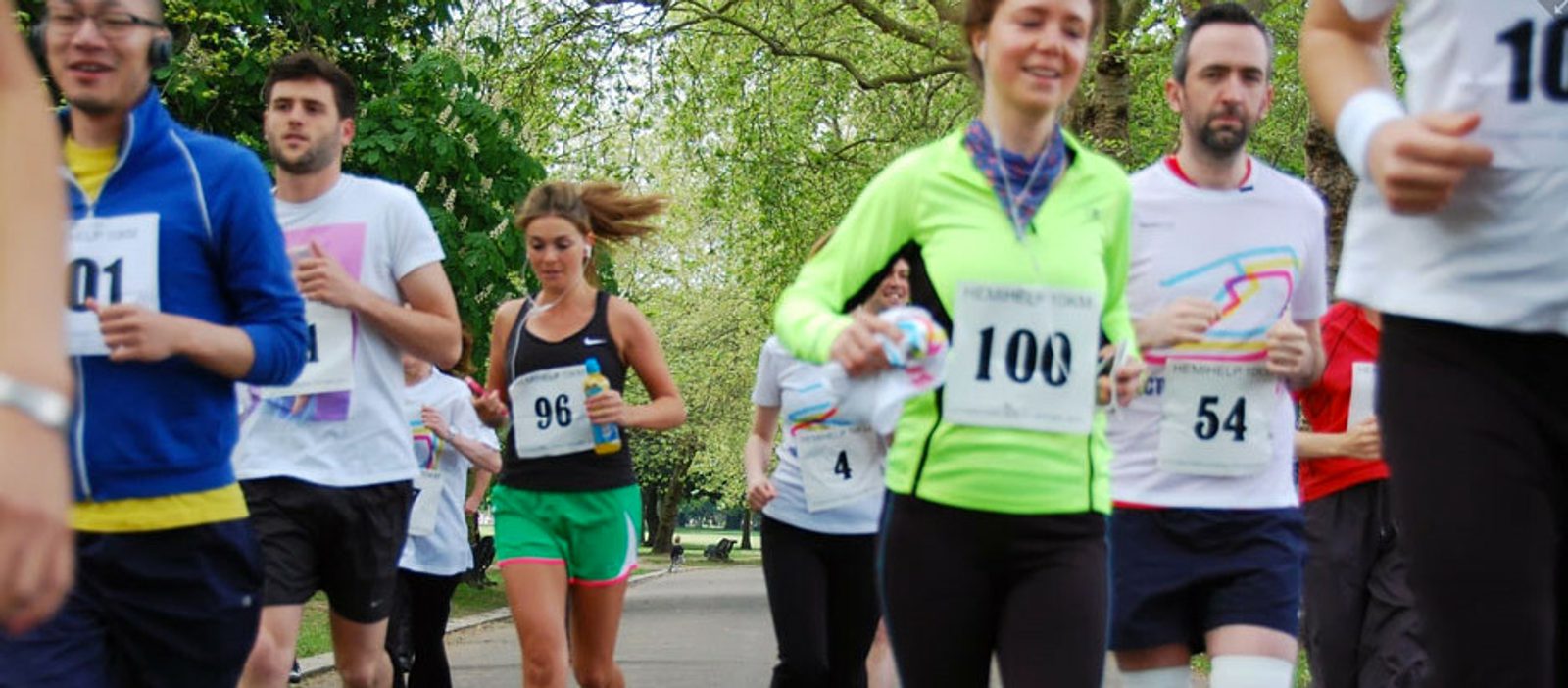 A group of people running in a race on a tree-lined path. They are wearing numbered bibs and athletic attire. Some runners are smiling and appear to be enjoying the event. The background shows lush green trees, indicating a park setting.