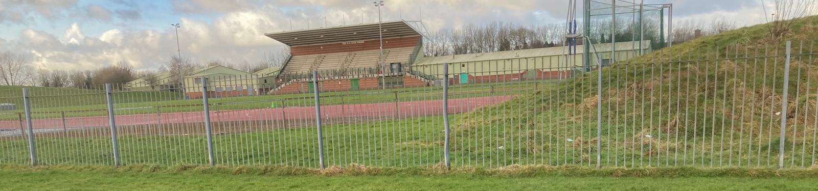 Panoramic view of an outdoor sports field with a track, grassy areas, metal fence in the foreground, and a grandstand with seating in the background. Cloudy sky is visible above.