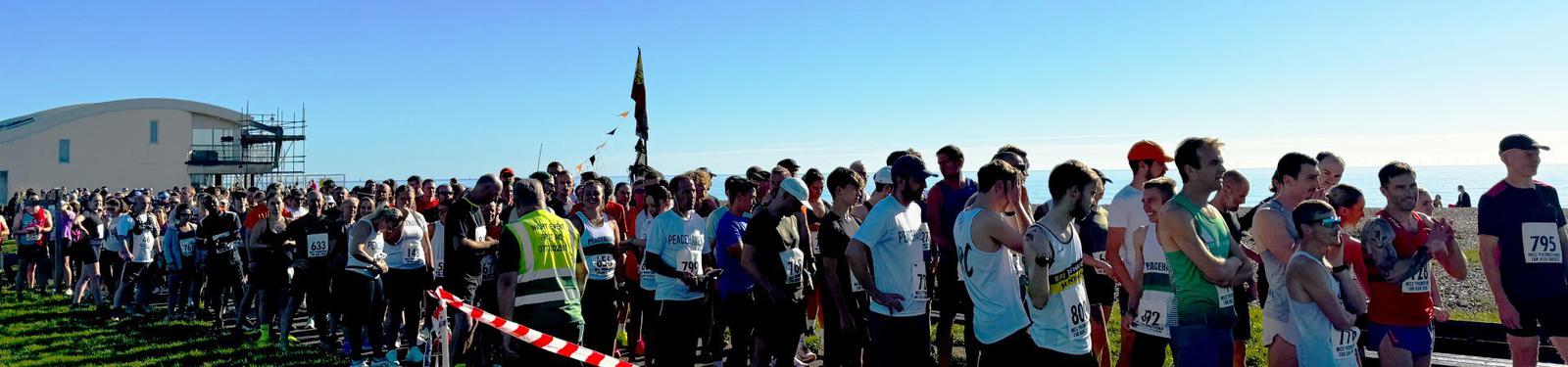 A large group of runners gathered at a starting line, wearing bib numbers and athletic gear. Some participants are adjusting their equipment or stretching. The sky is clear and blue, and there's a modern building in the background.