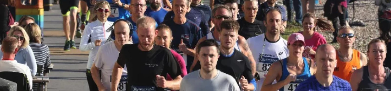 A group of runners is participating in a race on a sunny day. They are running on a paved path, with some onlookers and participants behind them. The runners vary in age and are wearing different athletic clothes, including numbered race bibs.