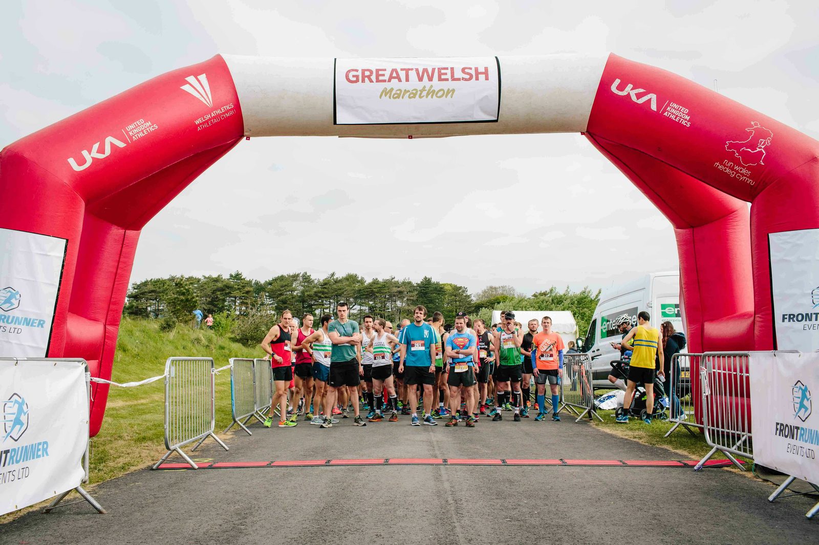 A group of runners stand at the starting line under a red and white inflatable arch that reads "Great Welsh Marathon." They are on a paved road with trees and grass in the background, preparing for the race to begin.