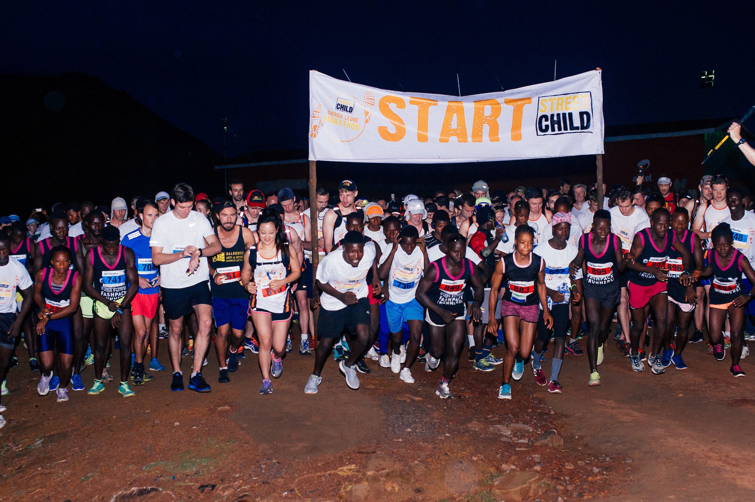 A diverse group of runners are starting a race at night under a banner that reads "START Street Child." They are concentrated and eager, with some wearing headlamps and race bibs. The background shows a dark sky and faint outlines of hills or buildings.