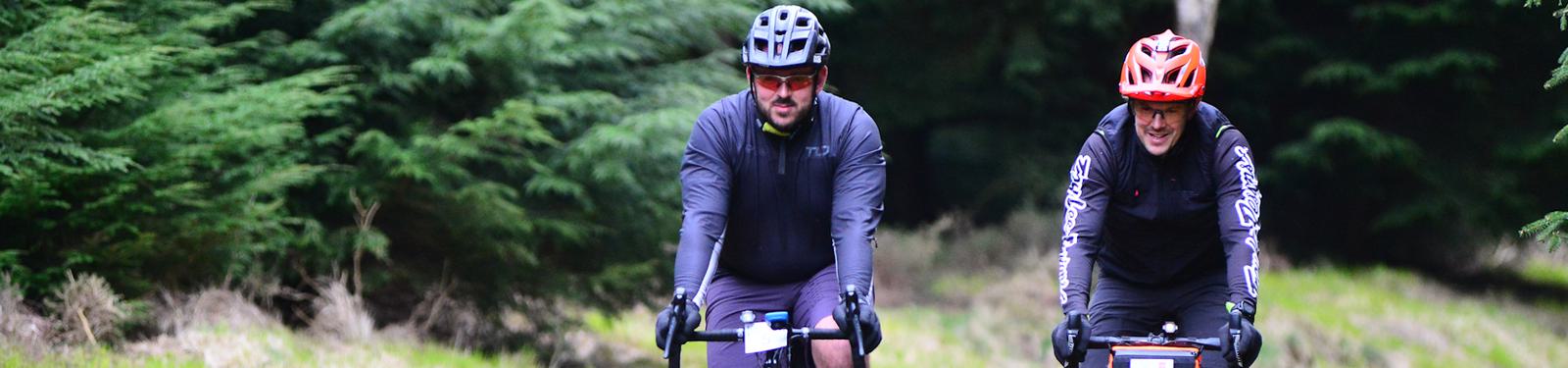 Two cyclists wearing helmets ride through a forested area. One cyclist wears a gray jacket and shorts; the other wears a red and black outfit. Both are on mountain bikes, surrounded by lush green trees.