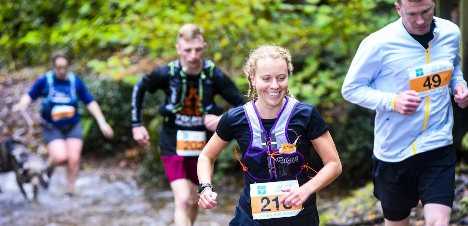 Four runners, three men and one woman, participate in a forest trail race. The woman in front is smiling while running. They all wear race bibs and athletic gear. The trail appears muddy and wet, with greenery in the background. A dog runs beside them.