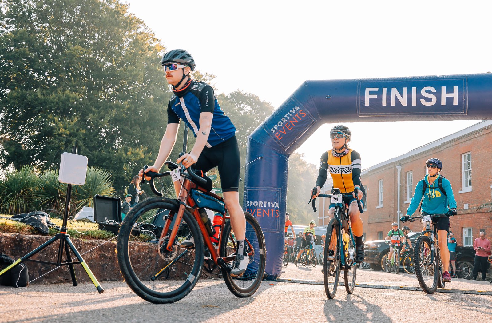 Cyclists crossing the finish line at a race event on a sunny day. An inflatable arch with the word "FINISH" is seen. One cyclist in blue leads, followed by another in a yellow jersey and a third in teal. Trees and buildings are in the background.