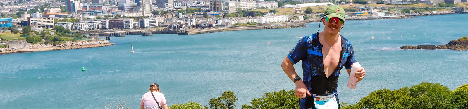A person in sports attire runs with a drink in hand along a scenic coastal path. In the background, there's a view of a city skyline across the water, with sailboats and rocky cliffs. Another person sits at the edge of the path.