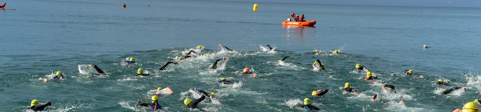 A group of swimmers wearing wetsuits and green swim caps race in open water with an orange inflatable boat and crew visible in the distance. Some swimmers in pink swim caps can be seen further ahead. The sky is clear and the water is slightly choppy.