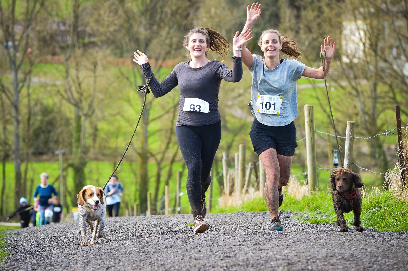 Two women jogging outdoors on a gravel path with their dogs. They are wearing race numbers and smiling with raised arms. The dogs, one larger and one smaller, trot happily beside them. Other runners can be seen in the background.