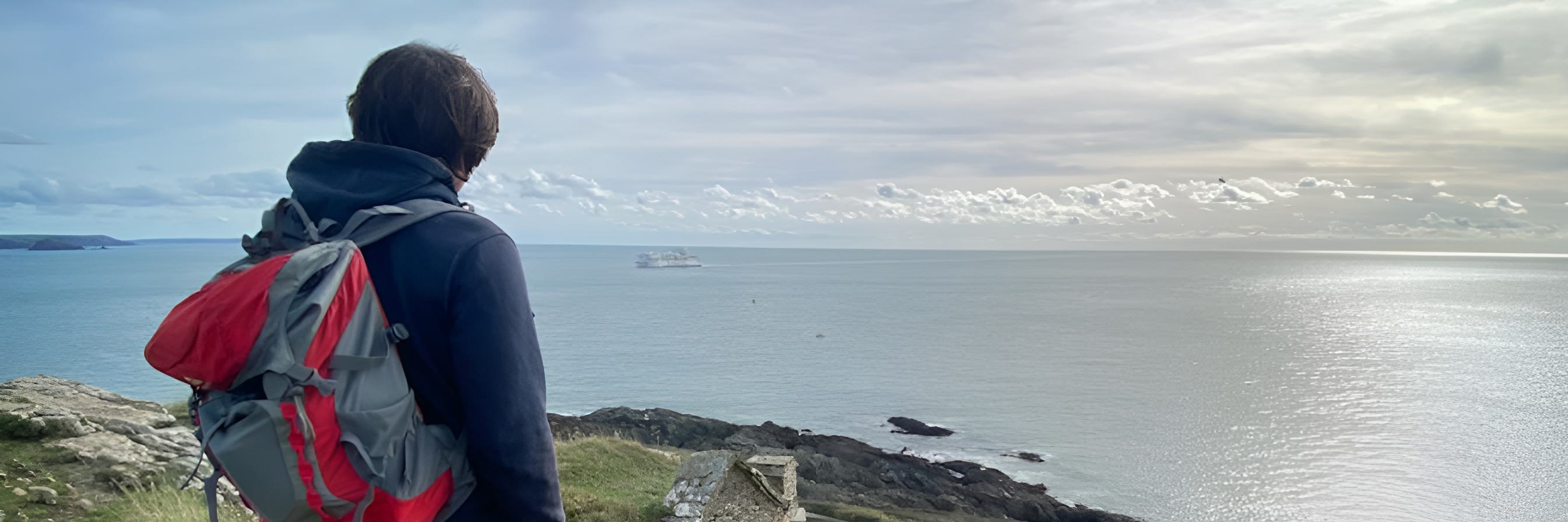 A person with a red and gray backpack stands on a grassy cliff, gazing at the calm sea. The sky is mostly cloudy with patches of sunlight reflecting on the water. A distant boat is visible on the horizon.