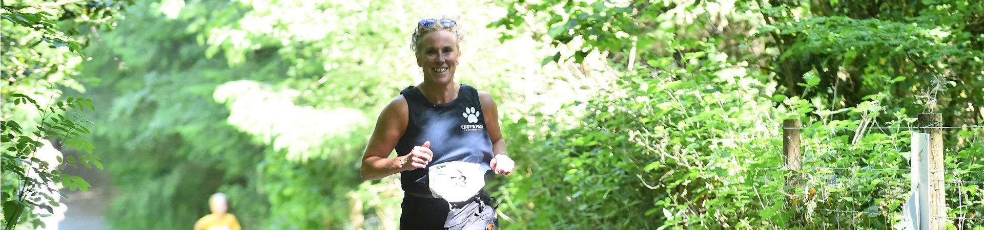 A woman runs along a sunlit, tree-lined path during a race, wearing a black tank top and race bib. Lush green foliage surrounds the trail and another runner is visible in the distant background.