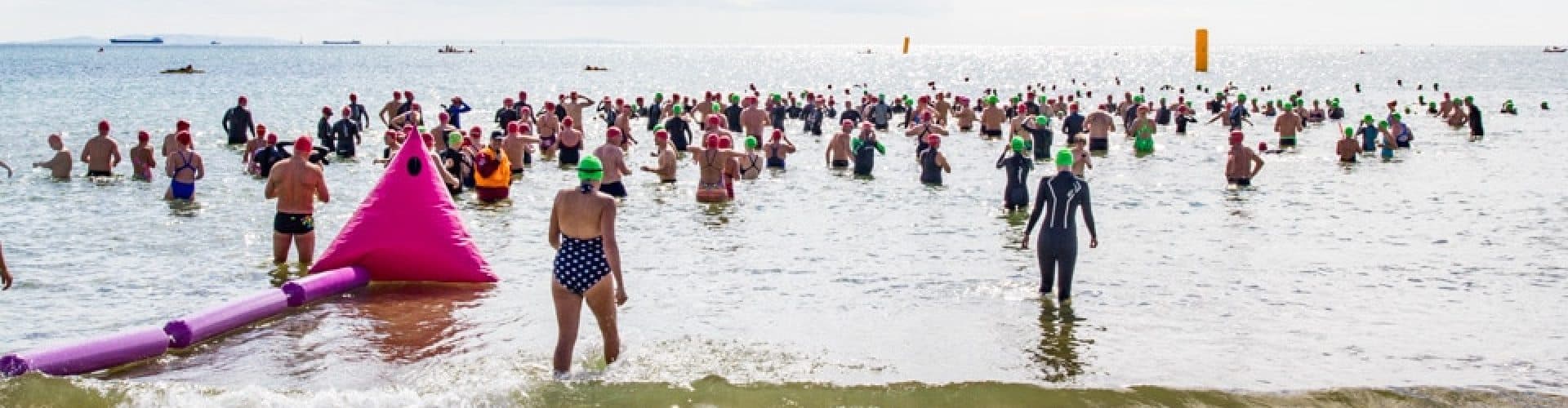 A large group of swimmers wearing colorful caps is gathered in shallow ocean waters near a beach, participating in an open-water swimming event. Inflatable buoys mark the course in the water.
