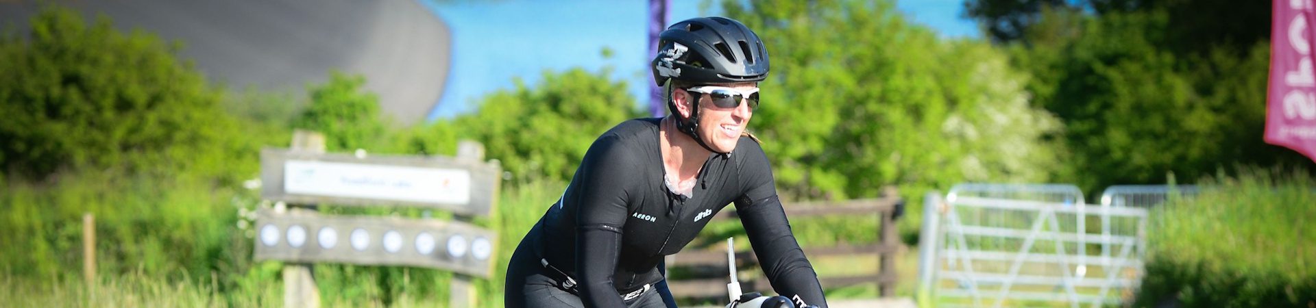 A cyclist wearing a black helmet, sunglasses, and a black outfit rides outdoors on a sunny day, surrounded by greenery and wooden fences.