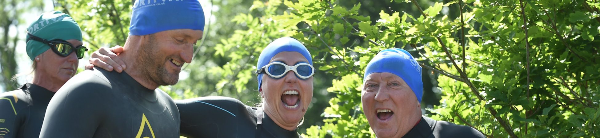 Four people in wetsuits and blue swim caps stand outdoors, smiling and laughing together, with greenery and sunlight in the background.