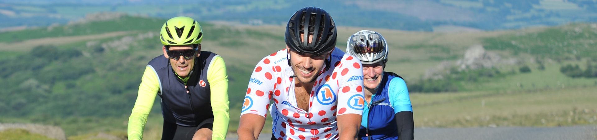 Three cyclists ride uphill on a rural road. The cyclist in front wears a white jersey with red polka dots and a black helmet, while the two behind wear bright yellow and blue gear. Green hills and countryside stretch out behind them.