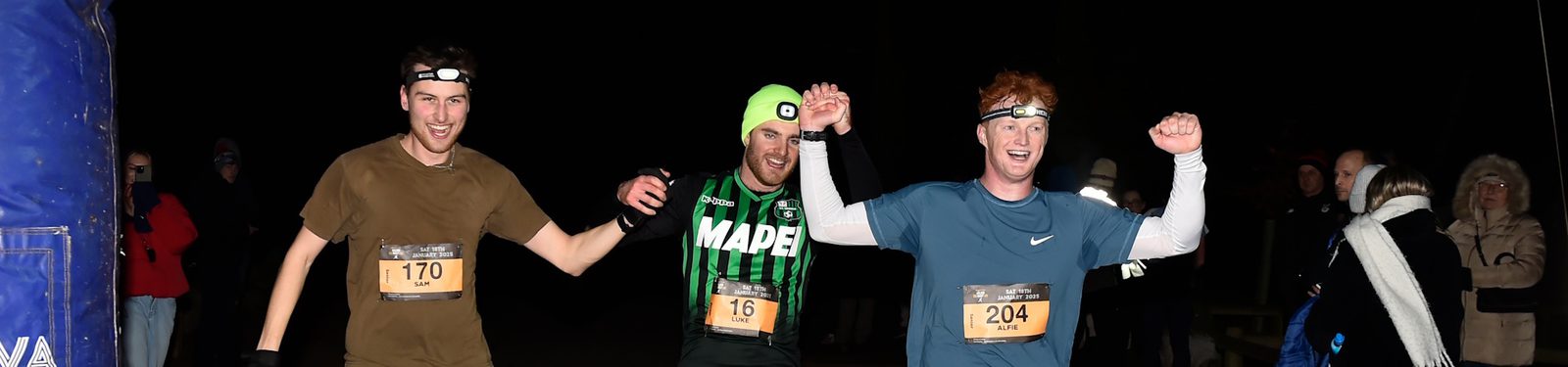 Three runners wearing headlamps and race bibs, two holding hands raised in celebration, smile as they cross a nighttime finish line. Spectators bundled in winter clothing watch in the background.