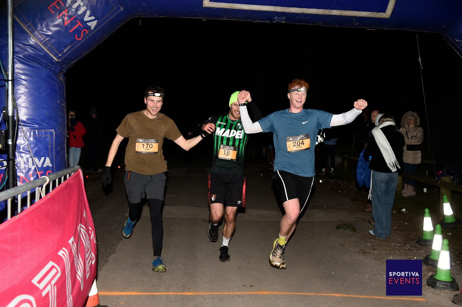Three male runners cross a nighttime race finish line together, holding hands in celebration. They wear numbered bibs and athletic gear, with cheering spectators and a “Sportiva Events” banner visible.