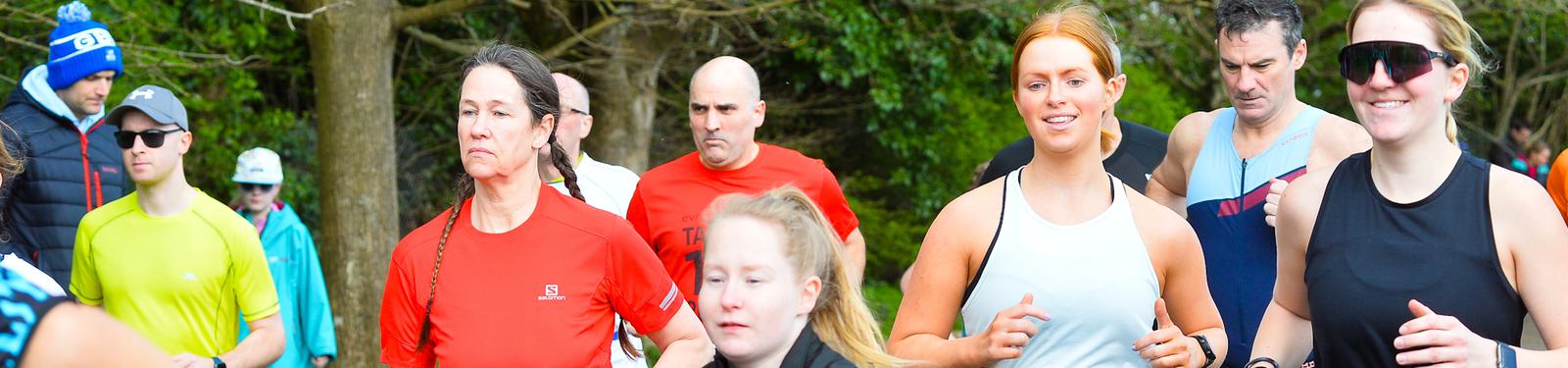 A group of people jogging outdoors. They are wearing athletic clothing, including colorful shirts and sunglasses. The background shows trees and some spectators. The mood is energetic and focused.