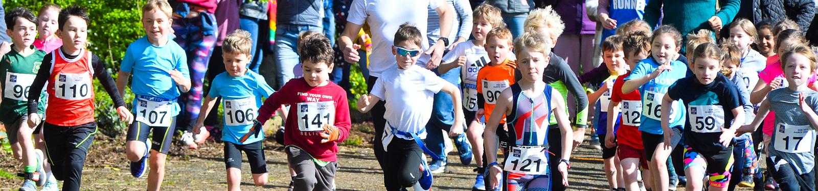 A group of children in athletic attire are energetically participating in a race, each wearing numbered bibs. The kids appear enthusiastic and are running on a sunlit path, with spectators in the background.
