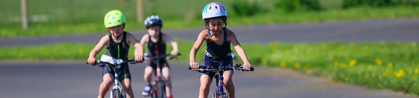 Three children wearing helmets are riding bicycles on a paved road in a green park. They appear to be participating in a cycling event, with the child in front concentrating on the road ahead.