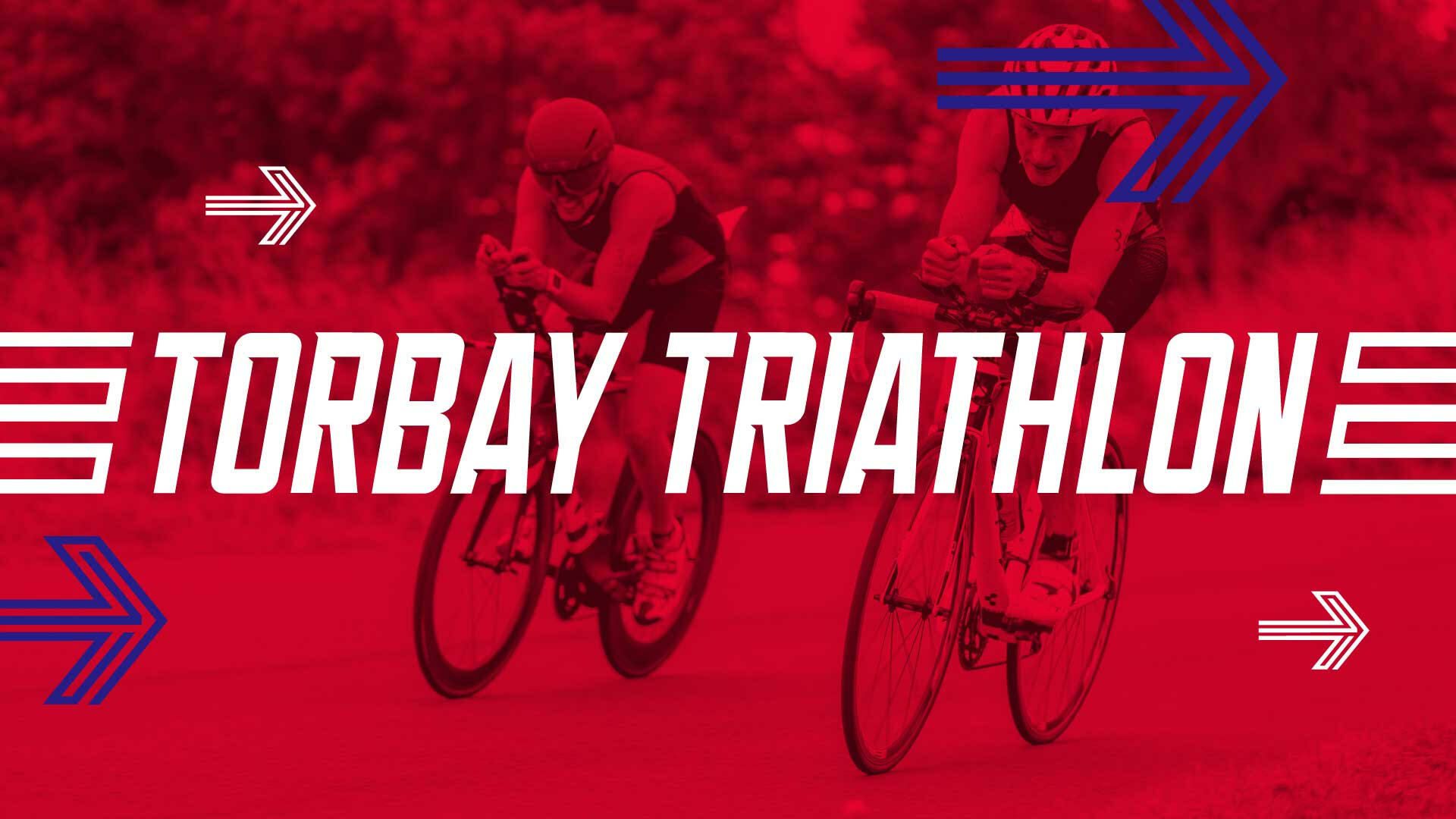 Two cyclists participating in the Torbay Triathlon ride on a road, both wearing helmets and racing gear. The image is overlaid with a red tint and the text "TORBAY TRIATHLON" in bold white letters, accompanied by decorative arrows in blue and white.