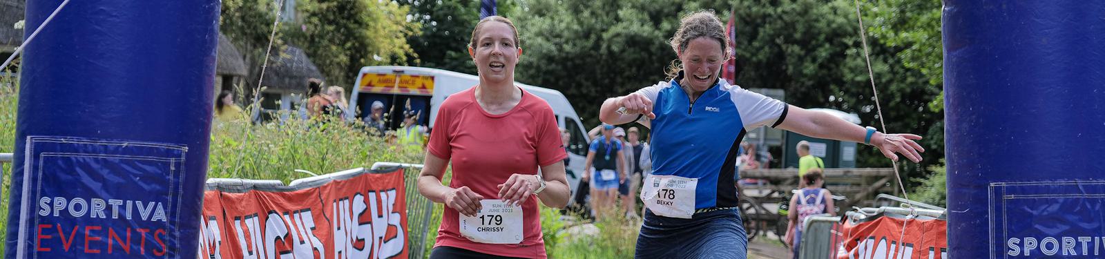 Two women are running a race, approaching the finish line together. They are wearing numbered bibs and smiling. Event signage and number banners are visible in the background, with trees and spectators nearby.