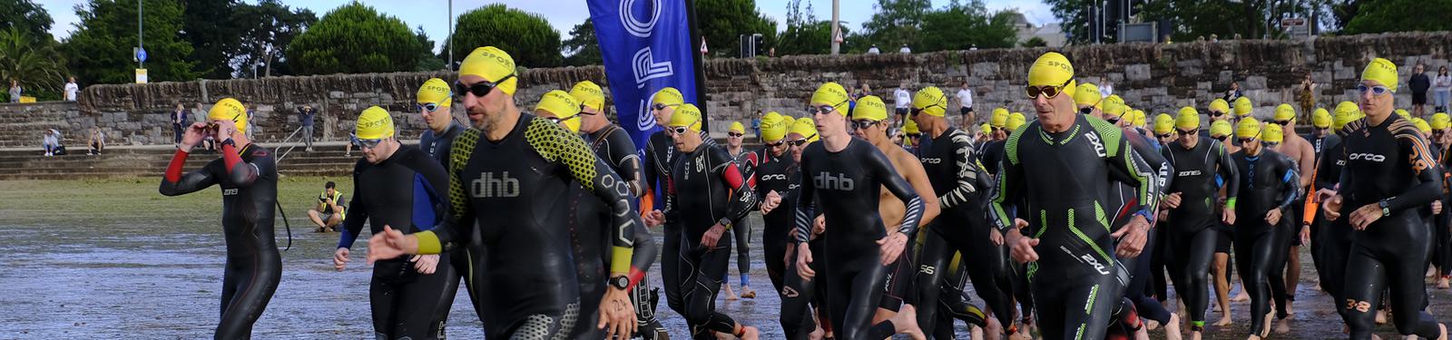 A group of athletes wearing wetsuits and yellow swim caps running along a beach. They appear to be participating in a triathlon, with a blue flag in the background. Trees and a stone wall are visible behind them.