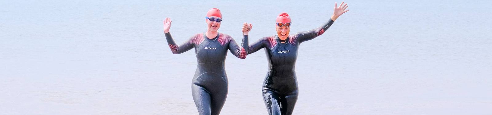 Two people wearing black wetsuits and red swim caps, with goggles, happily walking out of the water hand in hand, waving with their free hands. The background shows a calm, light blue sea.