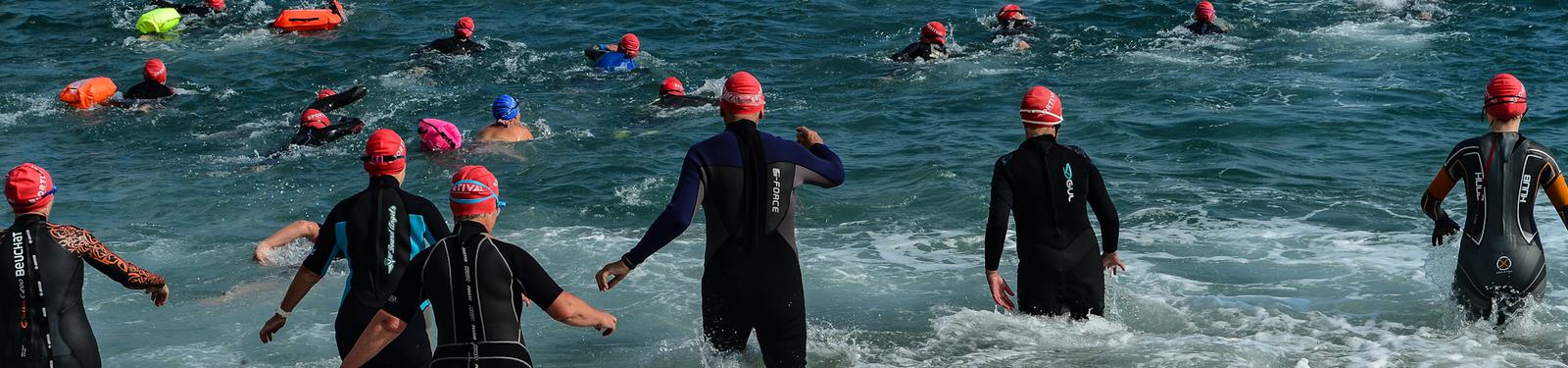 A group of swimmers wearing wetsuits and red swim caps enter the ocean, participating in an open-water race. The water is choppy, and some people are already swimming while others wade into the surf.