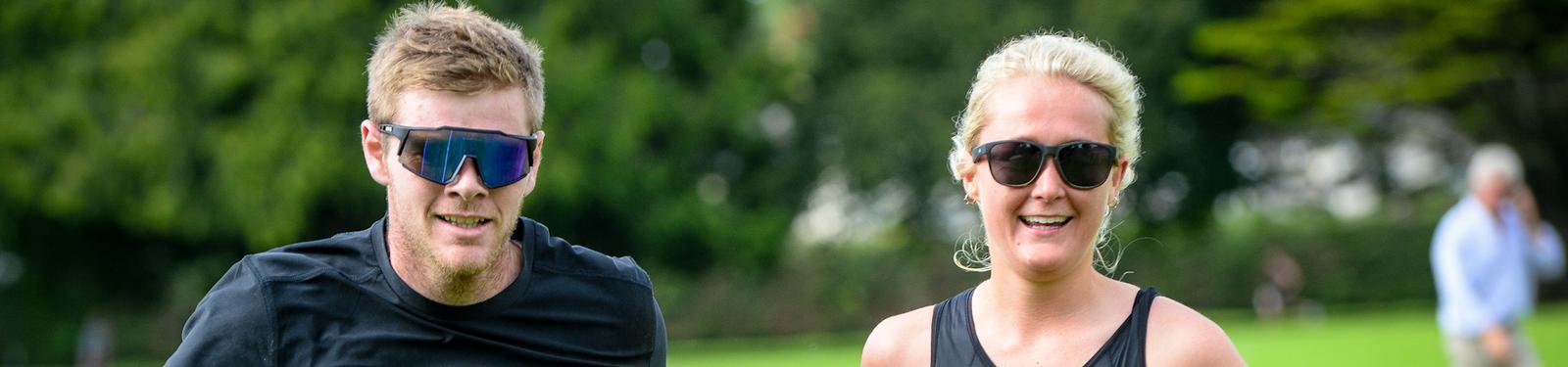 A man and a woman wearing sunglasses are jogging outdoors on a lush green field. Both are smiling and appear energetic, with trees and a person in the background.