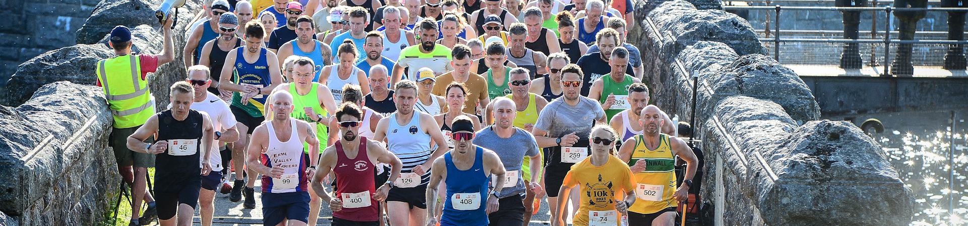 A large group of runners participating in a marathon on a narrow path between stone walls. They are wearing a variety of athletic clothing, some with numbered bibs. The sun is shining, and a person in a reflective vest is visible on the side.