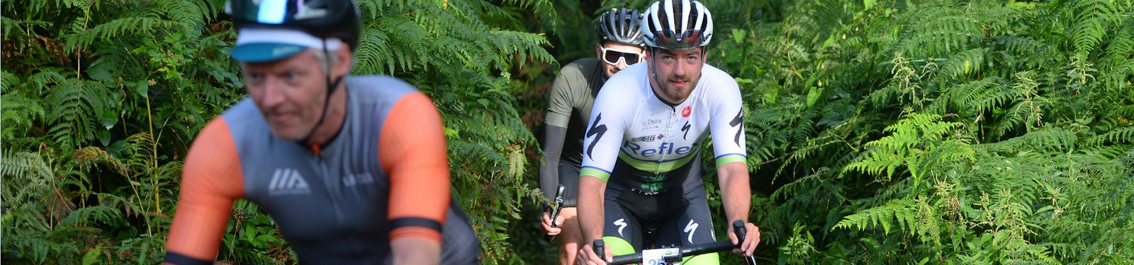 Three cyclists ride along a lush, green path surrounded by ferns. The focus is on a cyclist in white and green in the middle, wearing a helmet and smiling. The others are slightly out of focus, wearing orange and dark colors. The scene is vibrant and lively.