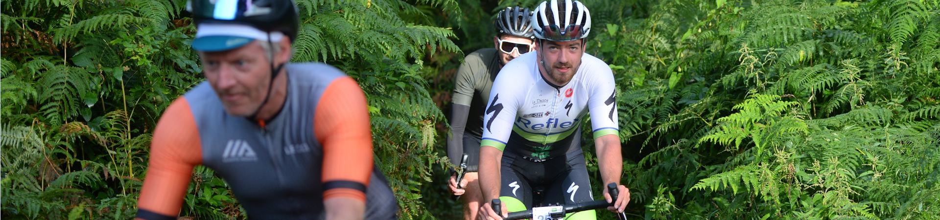 Three cyclists ride along a lush, green path surrounded by ferns. The focus is on a cyclist in white and green in the middle, wearing a helmet and smiling. The others are slightly out of focus, wearing orange and dark colors. The scene is vibrant and lively.