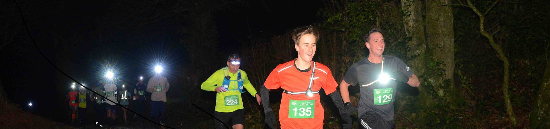 Runners participating in a nighttime race through a forest, wearing headlamps and athletic gear. The foreground shows two runners in red and black shirts, with illuminated bib numbers. Other runners and lights can be seen in the background.