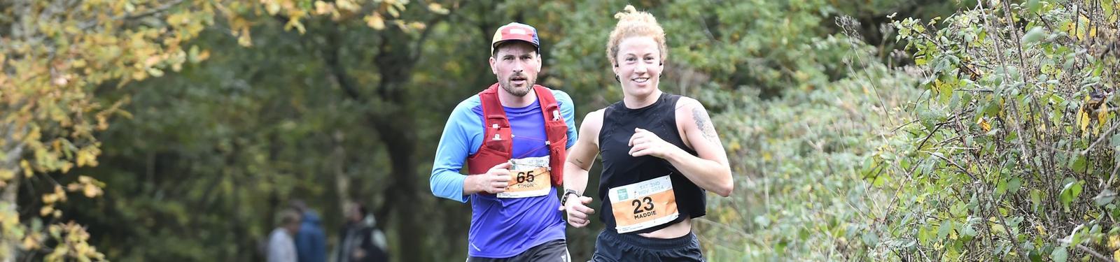 Two runners in a forest setting during a race. The woman in front wears a black tank top with a race bib numbered 23, while the man behind her wears a cap, blue long-sleeved shirt, and a bib numbered 65. Trees with green leaves are in the background.