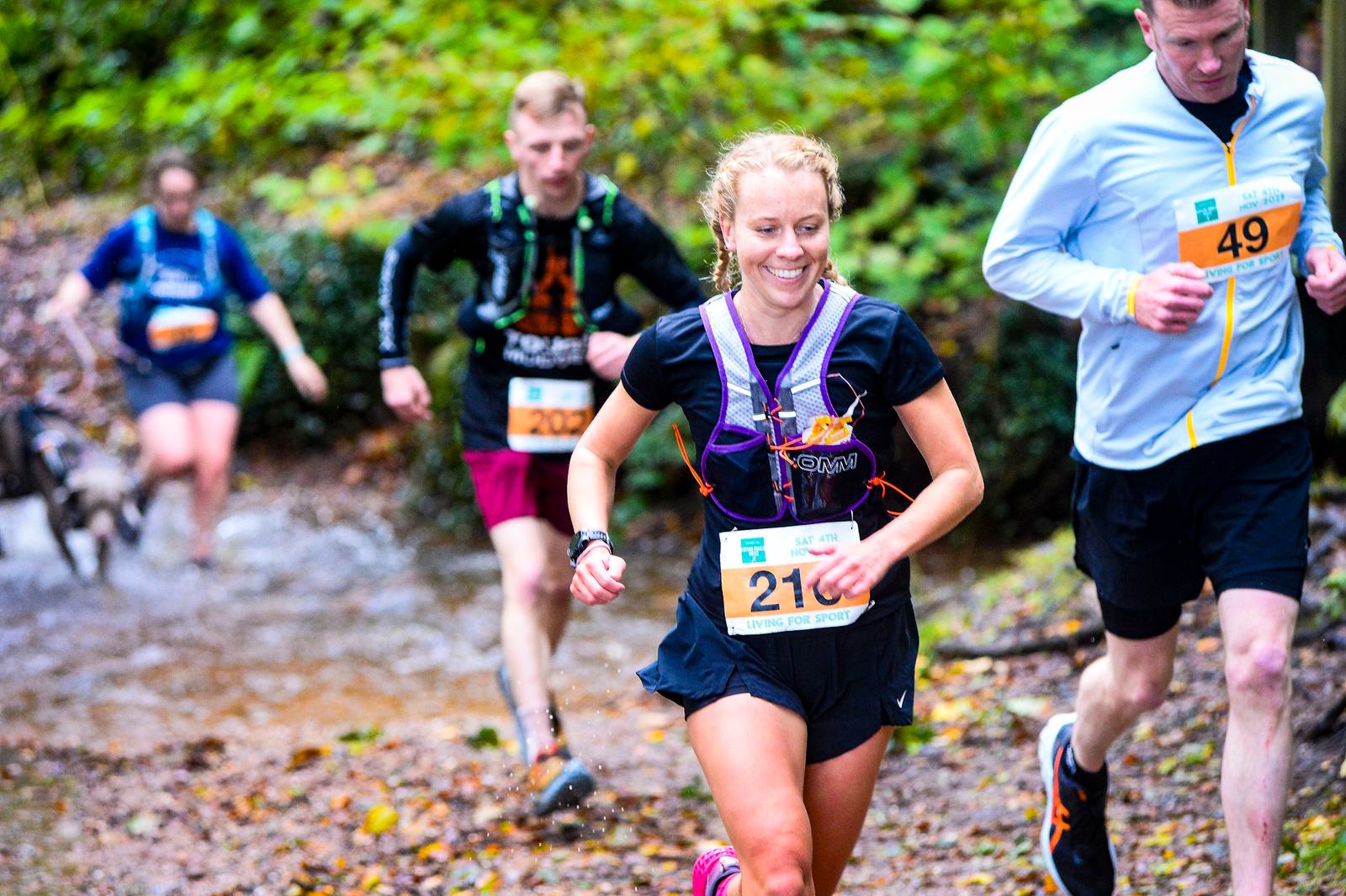 Runners navigate a muddy forest trail during a race. The foreground features a woman in a black shirt with a purple number bib smiling as she runs. Behind her, other participants jog along the wet path. Trees and fallen leaves surround them.