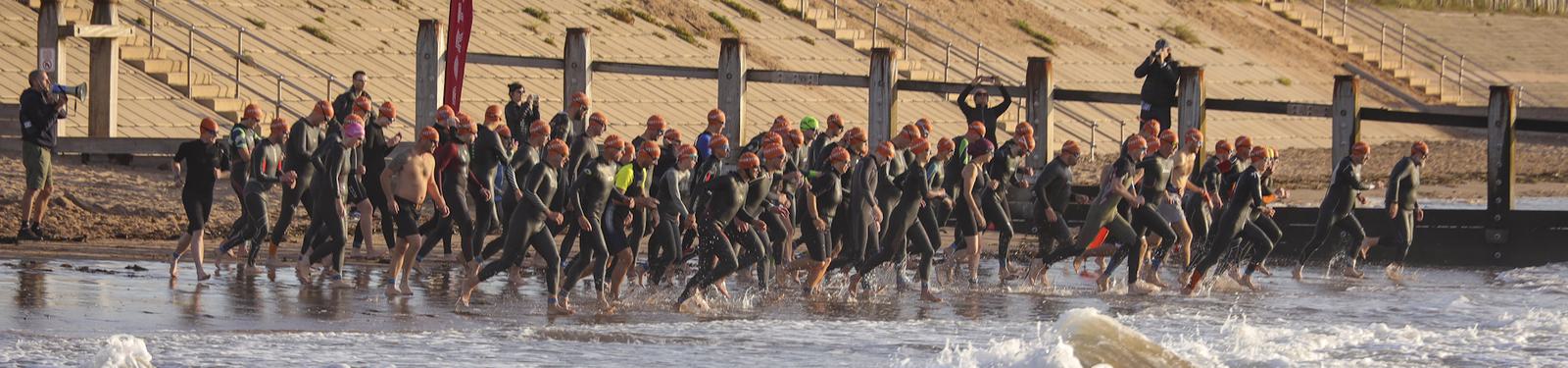 A group of triathletes wearing wetsuits and orange swim caps running from the beach into the ocean to start a race, with wooden posts and steps in the background.