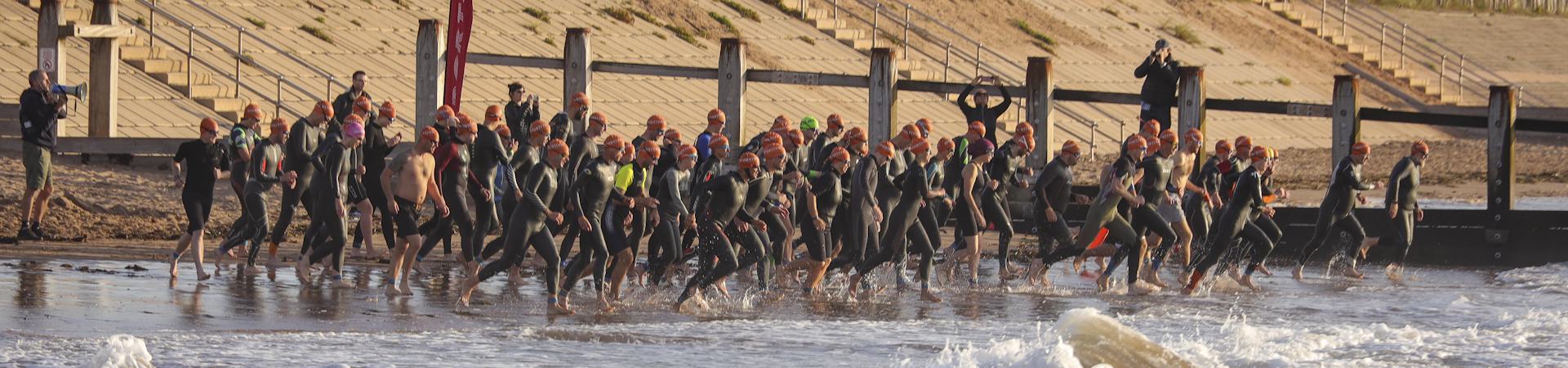 A group of triathletes wearing wetsuits and orange swim caps running from the beach into the ocean to start a race, with wooden posts and steps in the background.