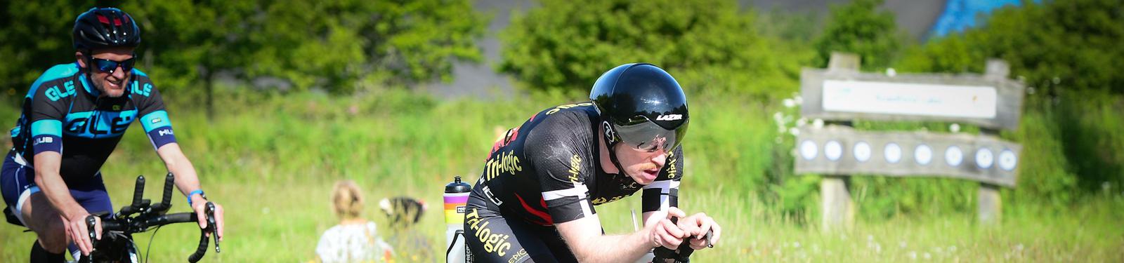 Two cyclists in racing gear ride on a sunny day through a green, grassy area. One cyclist is in a streamlined position wearing a black helmet. The background features a wooden sign and blurred foliage.