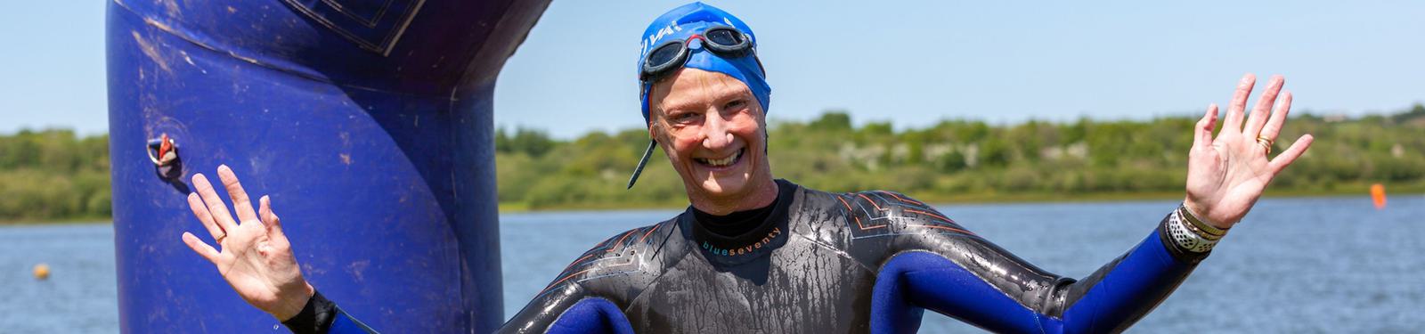 A person wearing a blue swim cap and black wetsuit stands near a large blue inflatable buoy, smiling with arms raised. They appear to be outdoors by a body of water under a clear sky.
