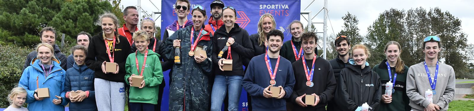 A group of people of various ages stand outdoors, smiling and holding medals or small boxes. They are in front of a banner that reads "Sportiva Events." Trees and a cloudy sky are visible in the background.