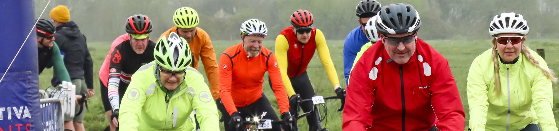A group of cyclists wearing colorful helmets and jackets participate in an outdoor cycling event. The background shows a misty landscape with green fields. They appear focused as they ride together on a cloudy day.