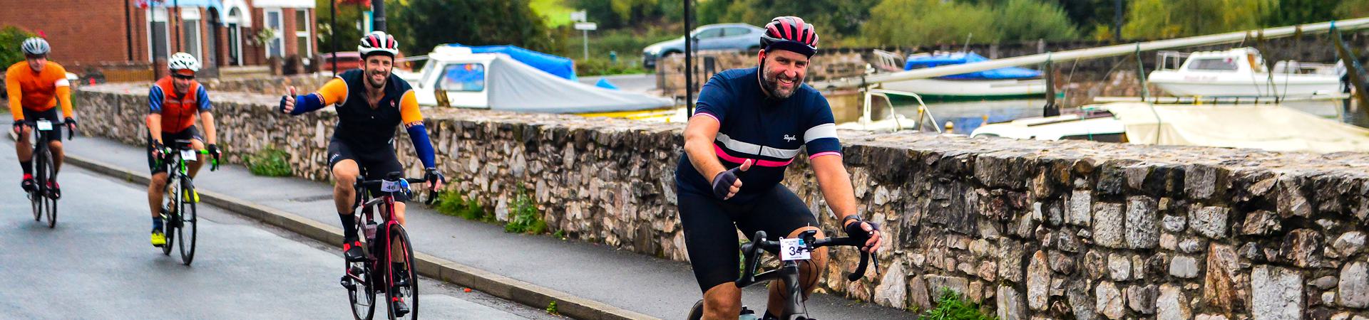 Cyclists ride along a riverside path beside a stone wall, with moored boats visible in the background. The lead cyclist smiles and gestures with a thumbs-up, while others follow closely behind. The scene is vibrant and energetic.