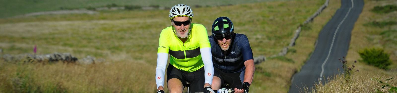 Two cyclists wearing helmets and sunglasses ride along a rural road surrounded by grassy fields. They appear to be in a race or organized cycling event, enjoying a sunny day.