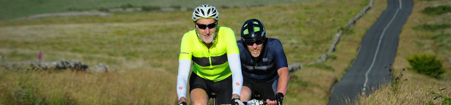 Two cyclists wearing helmets and sunglasses ride along a rural road surrounded by grassy fields. They appear to be in a race or organized cycling event, enjoying a sunny day.