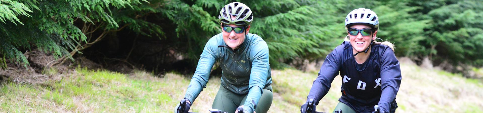 Two people wearing helmets and sunglasses are riding bicycles on a grassy path. Both are smiling, enjoying the outdoor ride. Lush green trees line the path, creating a natural background.