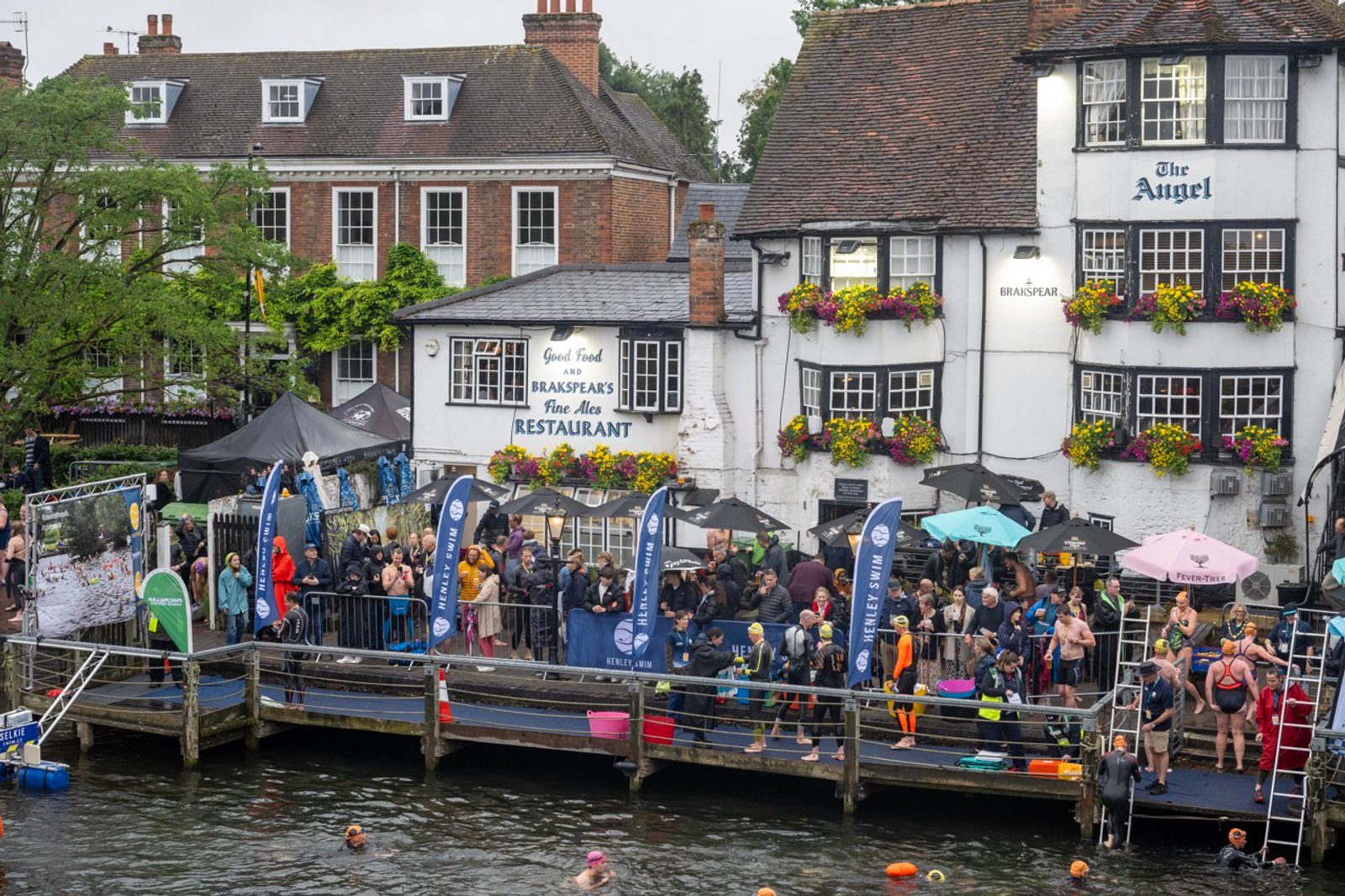 A bustling riverside scene with people gathering on a dock near the buildings of a restaurant and hotel adorned with flowers. Participants appear to be preparing for a swimming event, with some already swimming in the river and colorful umbrellas providing shelter.