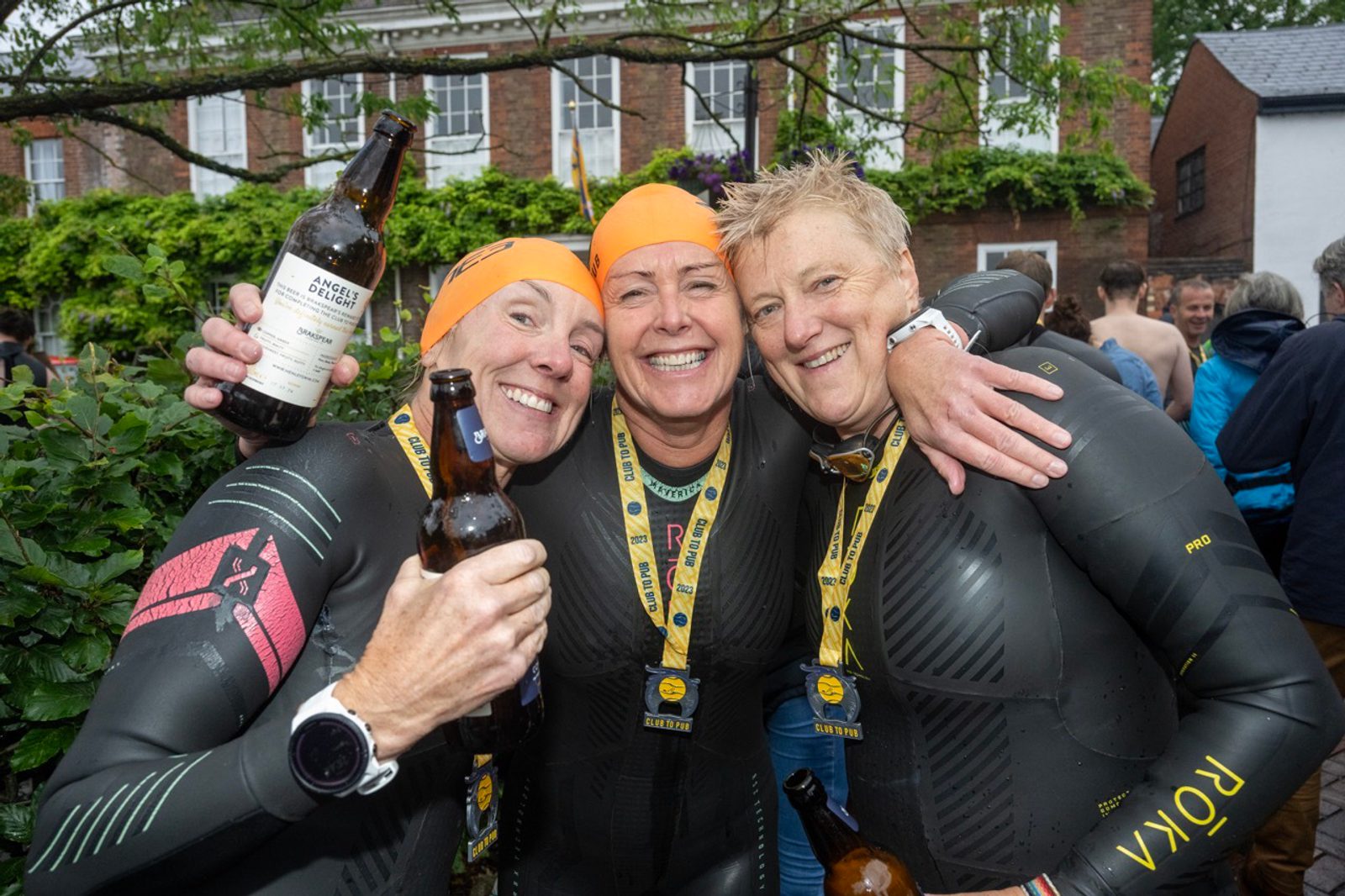 Three people in wetsuits and orange swim caps, each holding a beer bottle, are smiling and embracing while posing for a photo. They are wearing medals around their necks and seem to be celebrating their achievement. A crowd and buildings are visible in the background.