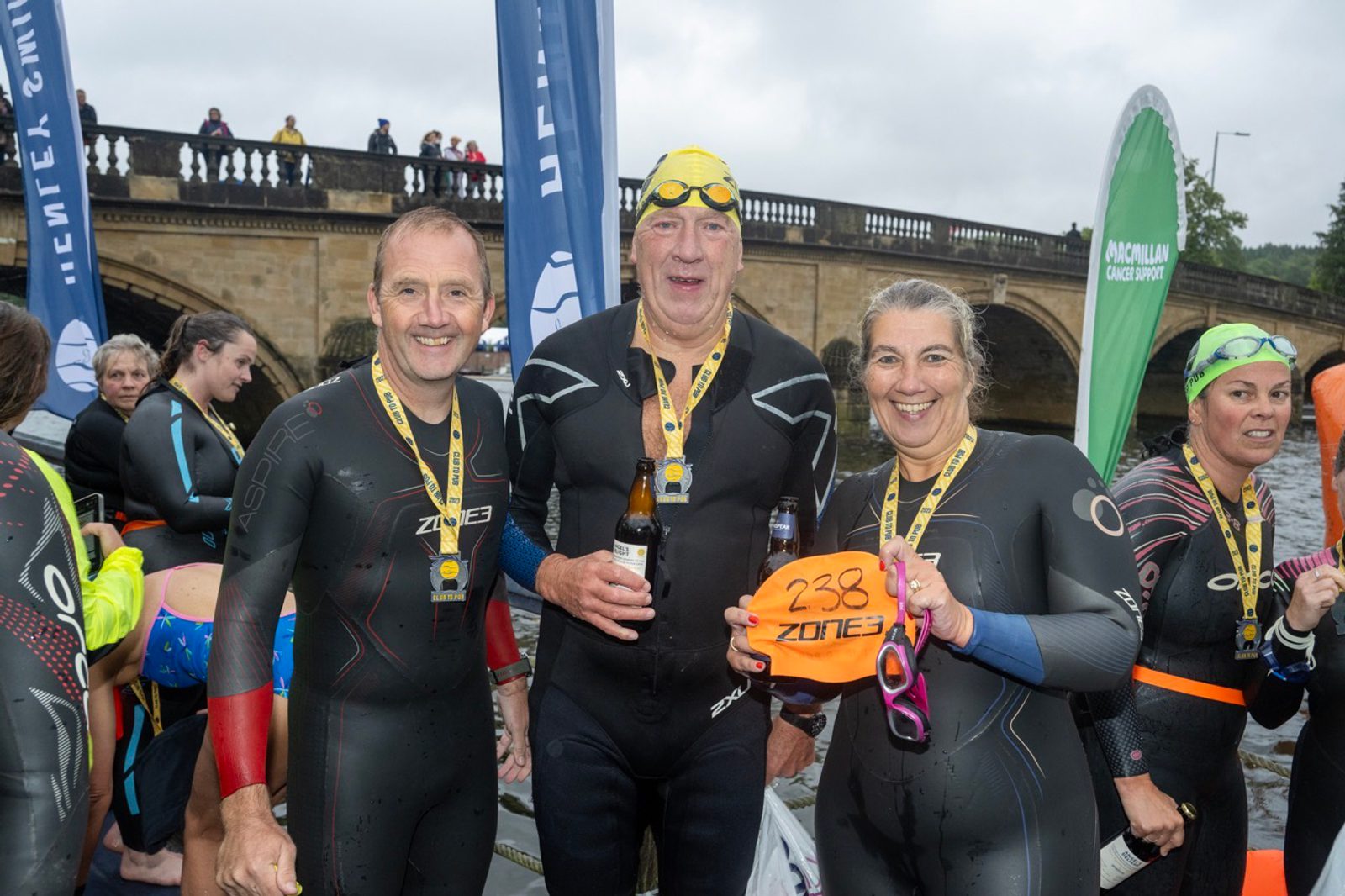 Participants in wetsuits, wearing medals, pose smiling after a swim race. One holds an orange buoy, another a beer, with a bridge and spectators in the background. Blue and green event banners are visible, and the weather appears cloudy.
