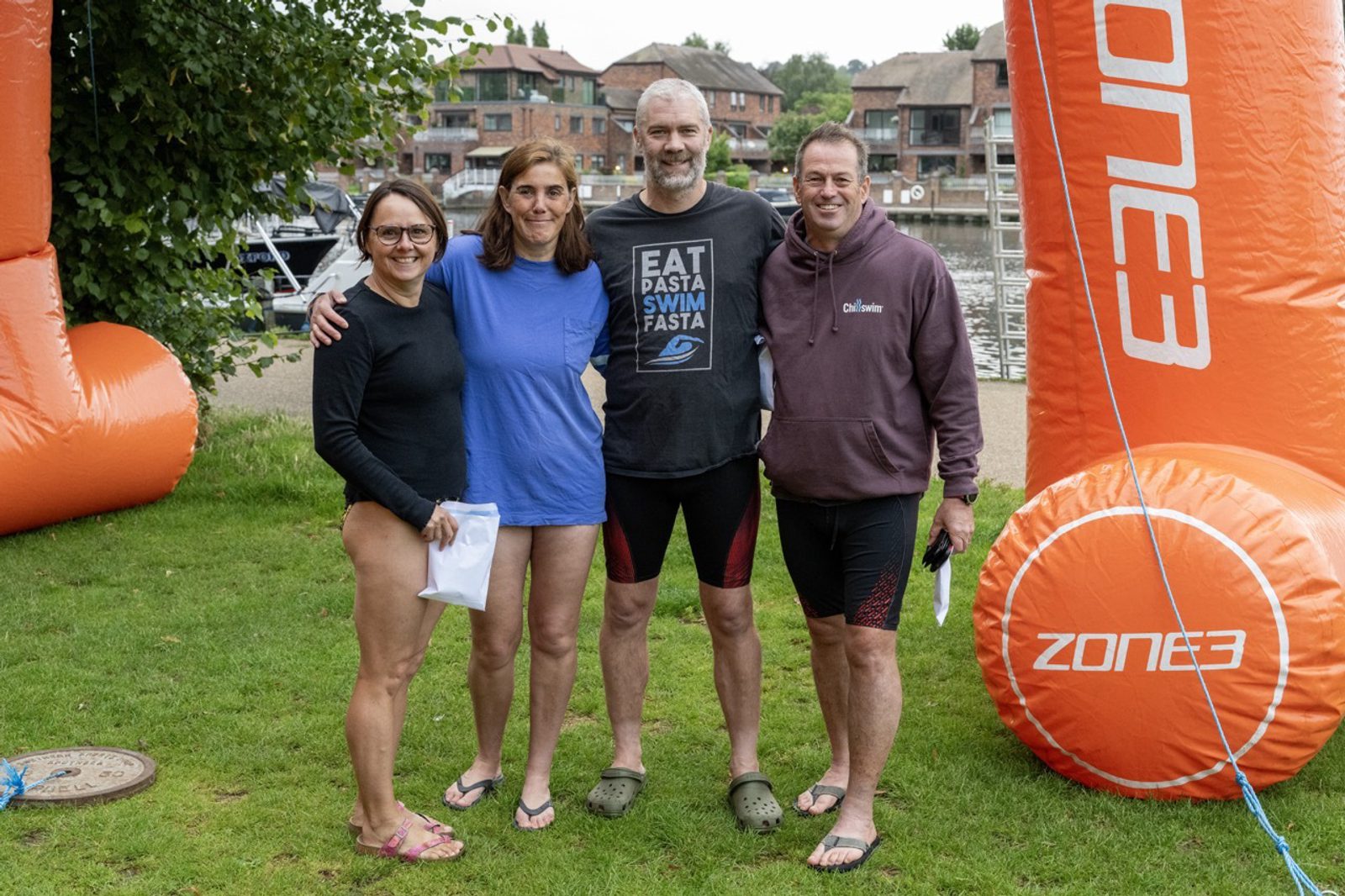 Four people pose together in front of orange inflatable markers labeled "ZONE3" at an outdoor event. They are standing on grass near a body of water with buildings in the background. Two women are on the left, wearing swimsuits, and two men are on the right, in athletic wear.